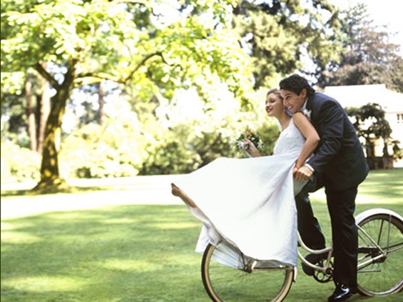 Groom riding bicycle, bride sitting on handlebars, side view